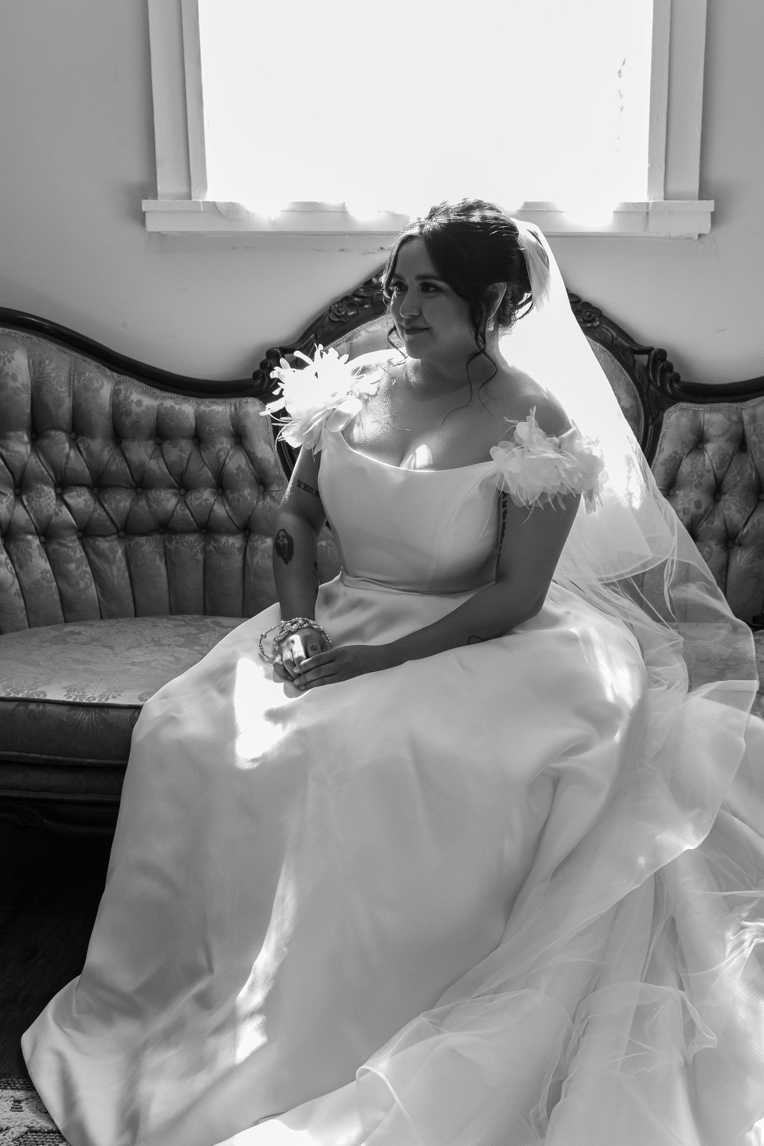 A bride in a white wedding dress reclines elegantly on a tufted couch in a black and white photograph.