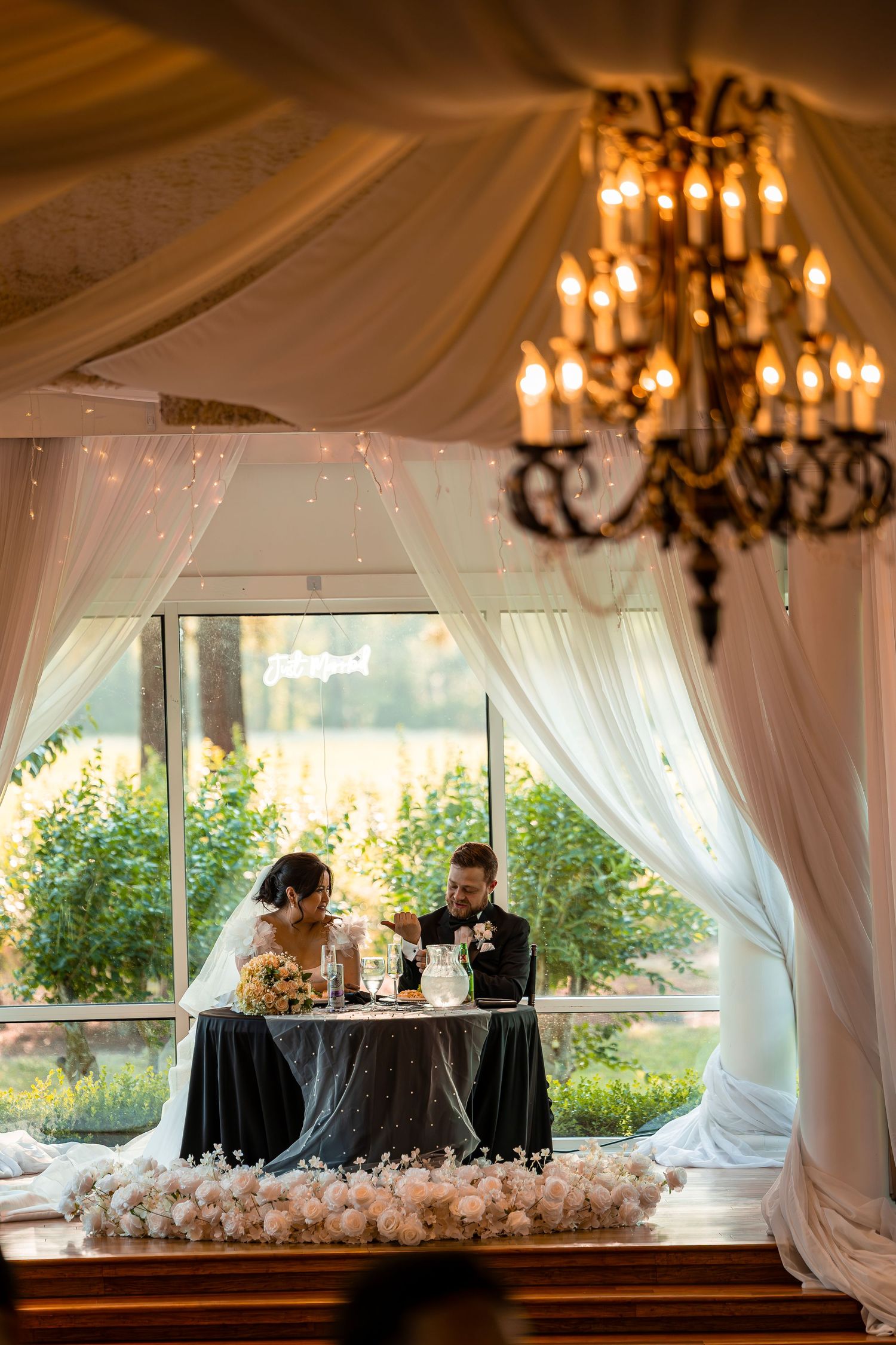 Elegant wedding reception setup in a white draped tent with chandeliers and decorative table settings.