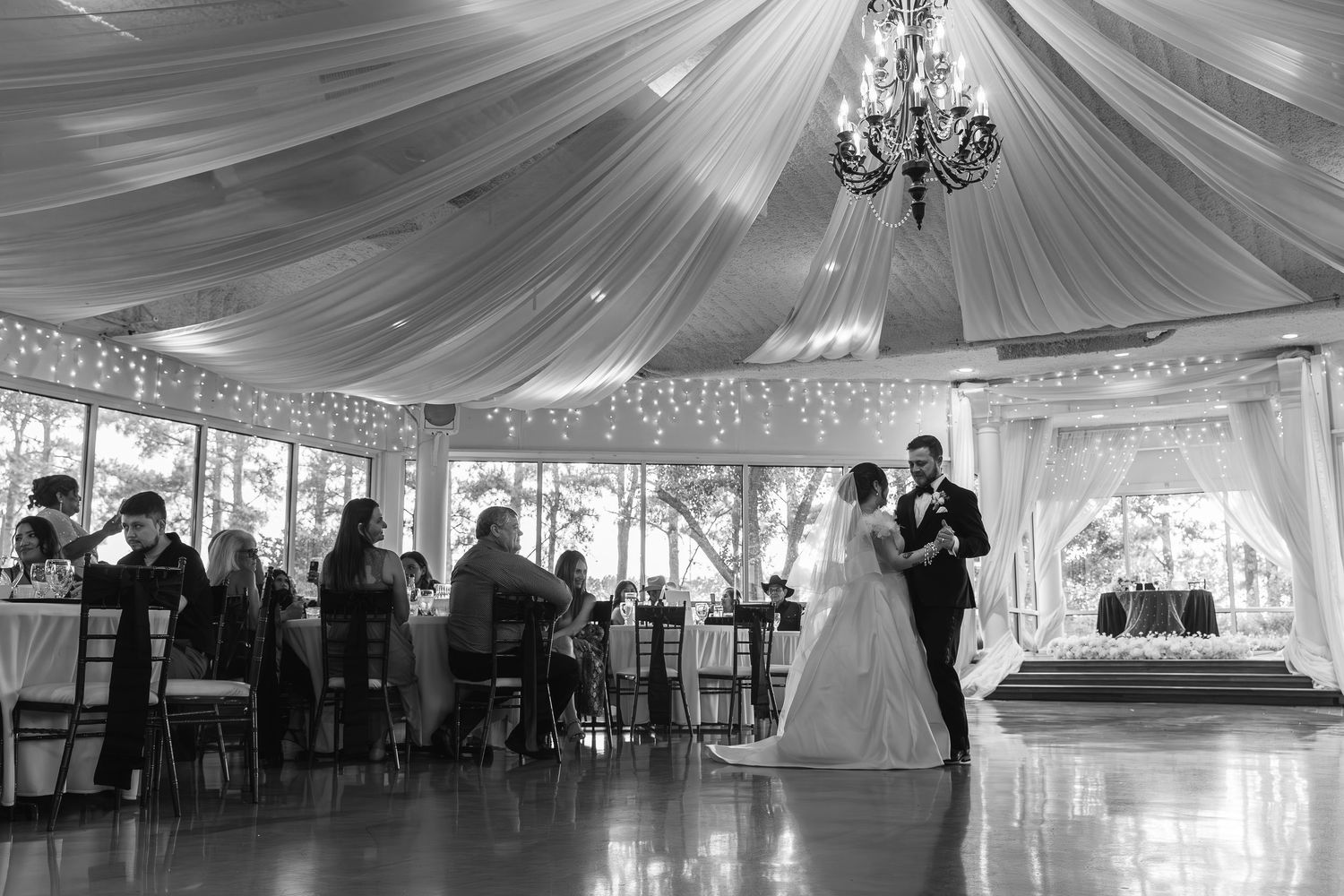 Wedding reception venue decorated with elegant white fabric draping and crystal chandeliers in a spacious ballroom.
