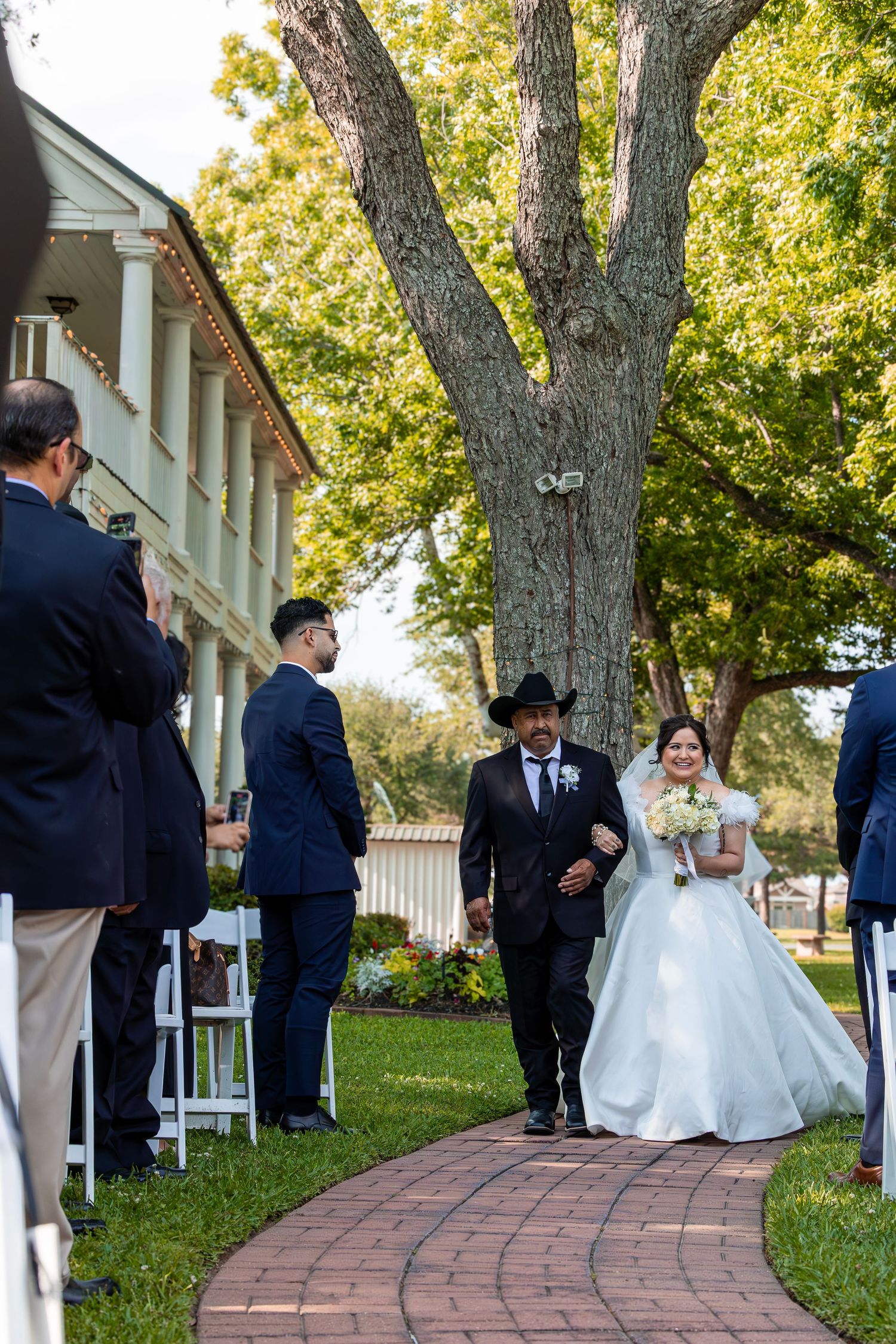 Wedding ceremony takes place outdoors on brick pathway with white chairs and tall trees providing natural shade.