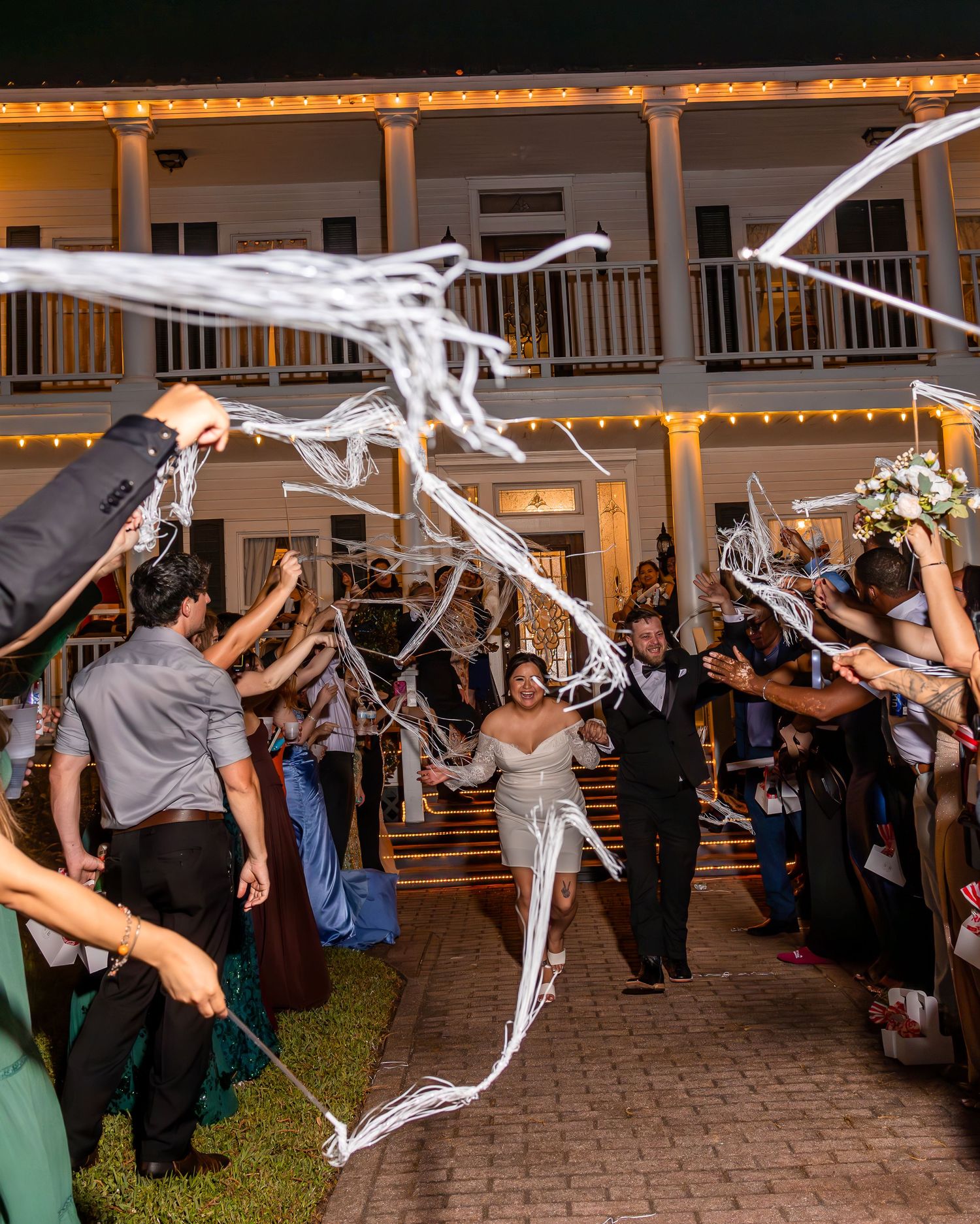 Wedding guests wave white streamers in celebration during an outdoor evening reception at a venue.