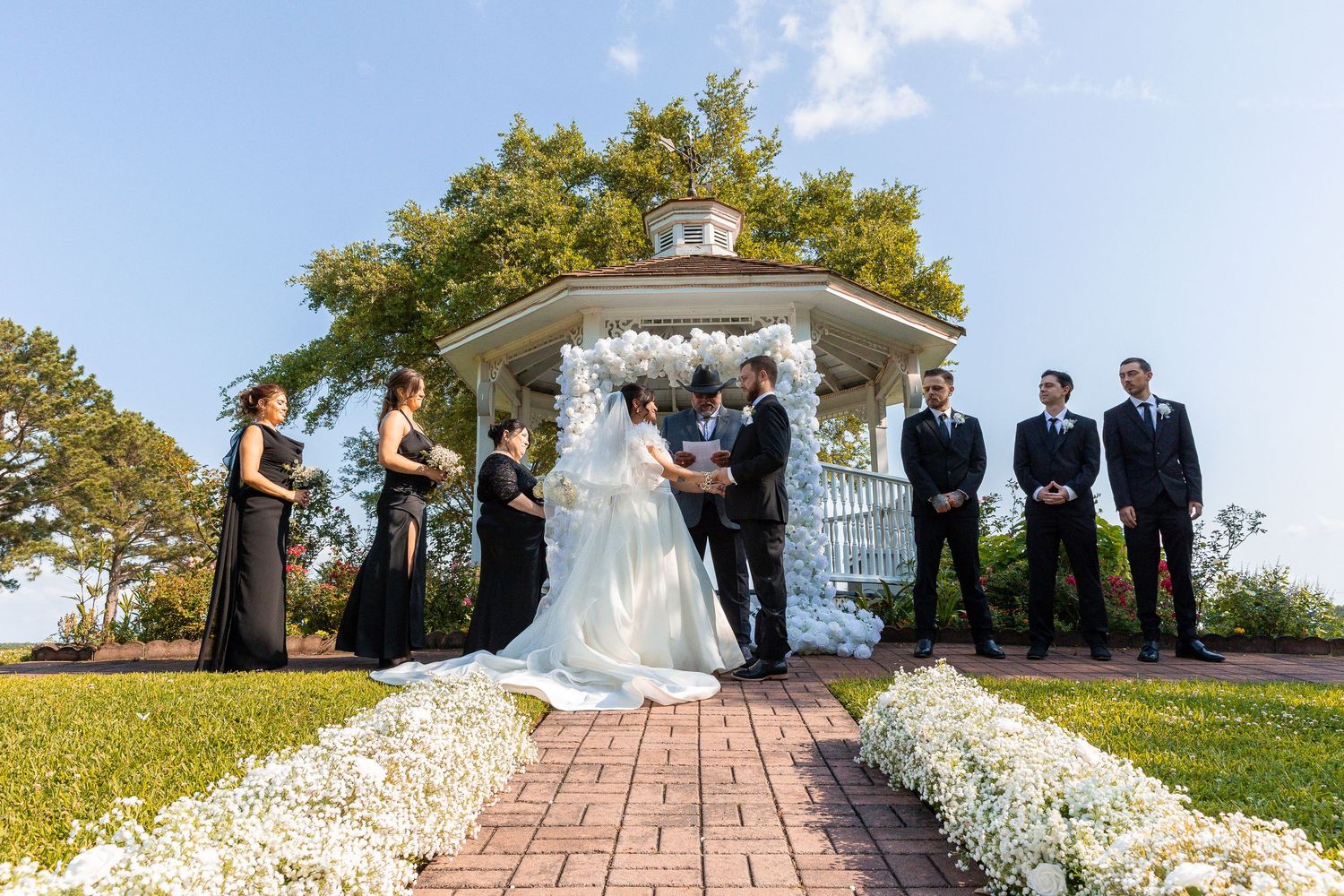 Wedding party stands on brick pathway surrounded by white flower petals and greenery on sunny day.