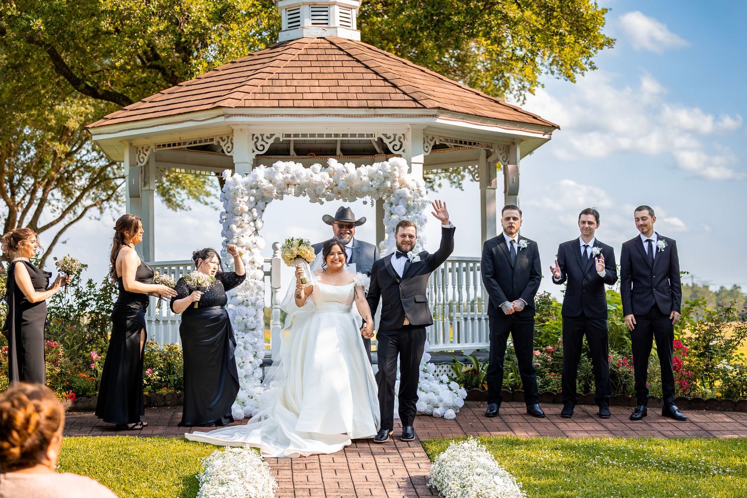Wedding party poses outdoors in front of a white gazebo on a sunny autumn day with colorful trees in the background.