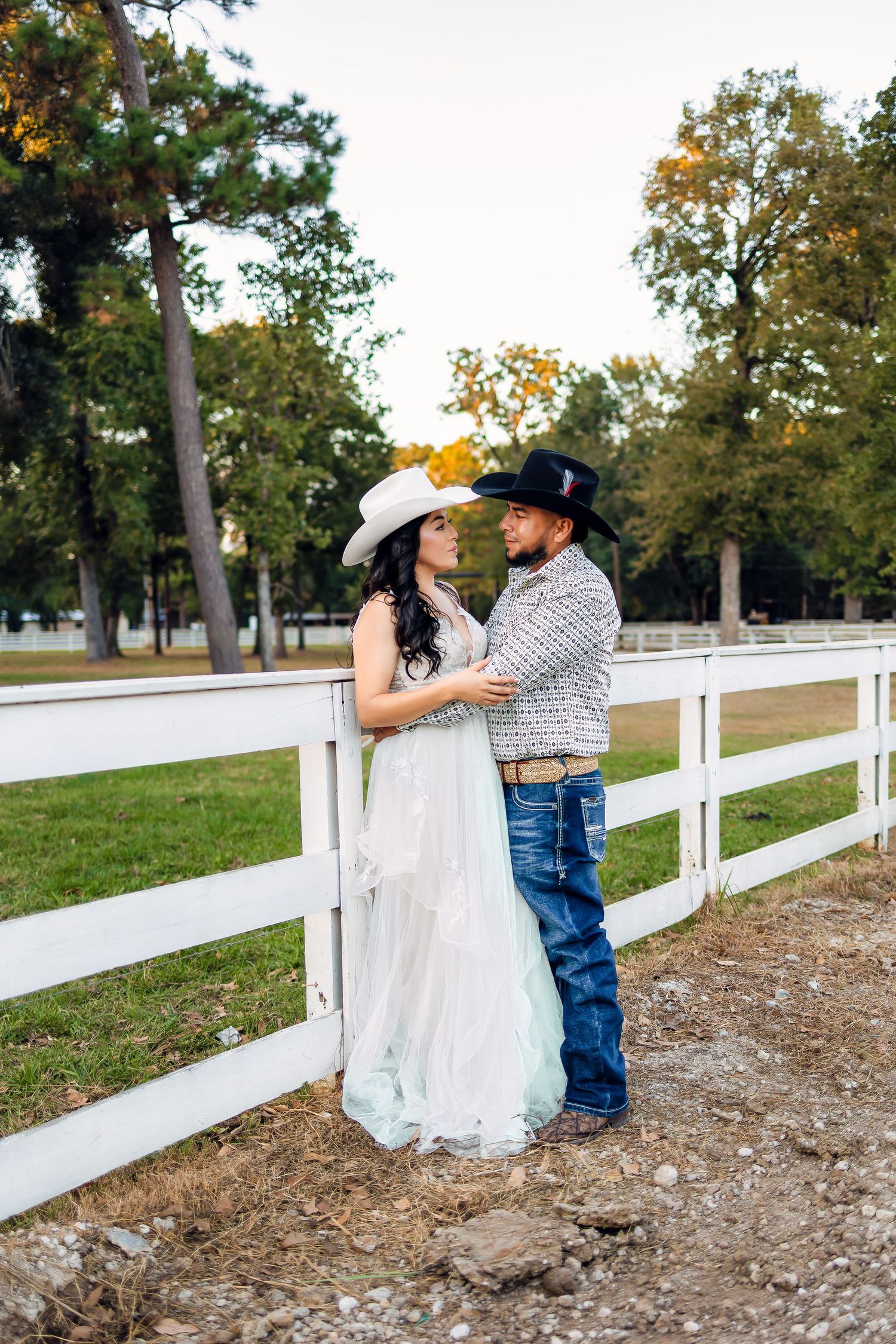 A couple shares a romantic moment along a white fence during a rustic countryside photo session.