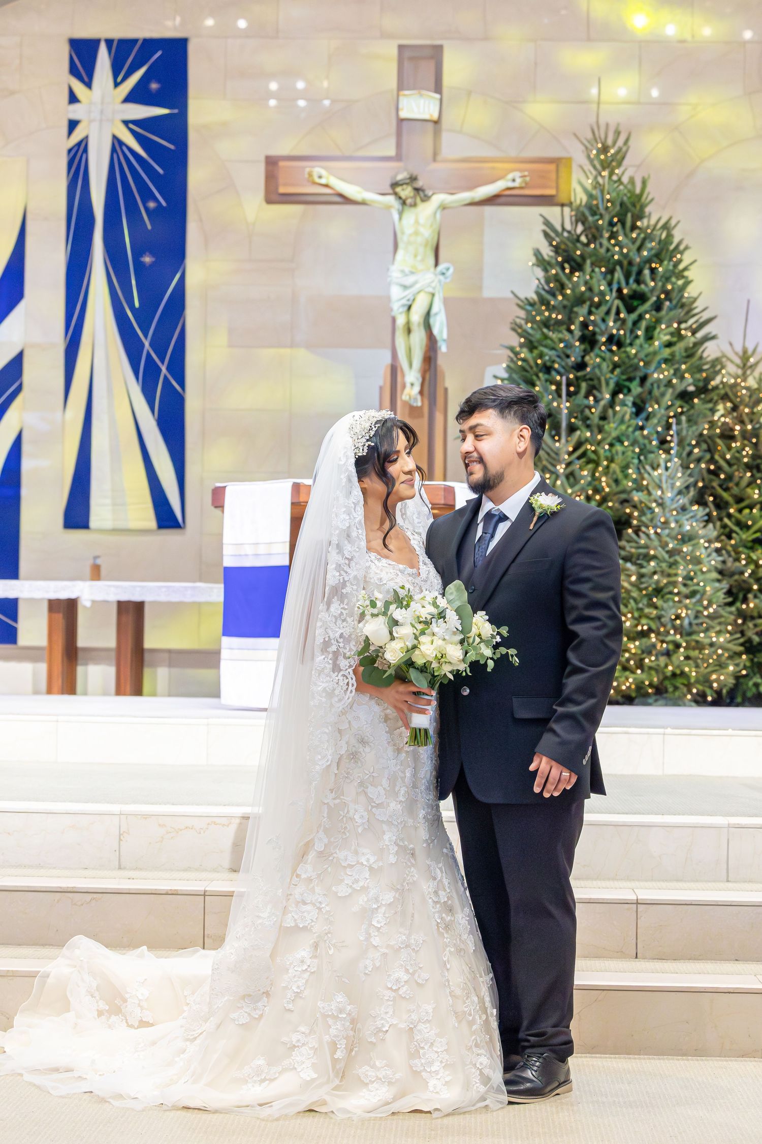 A bride and groom stand together at the altar in a church with blue and white decorative banners and potted trees.