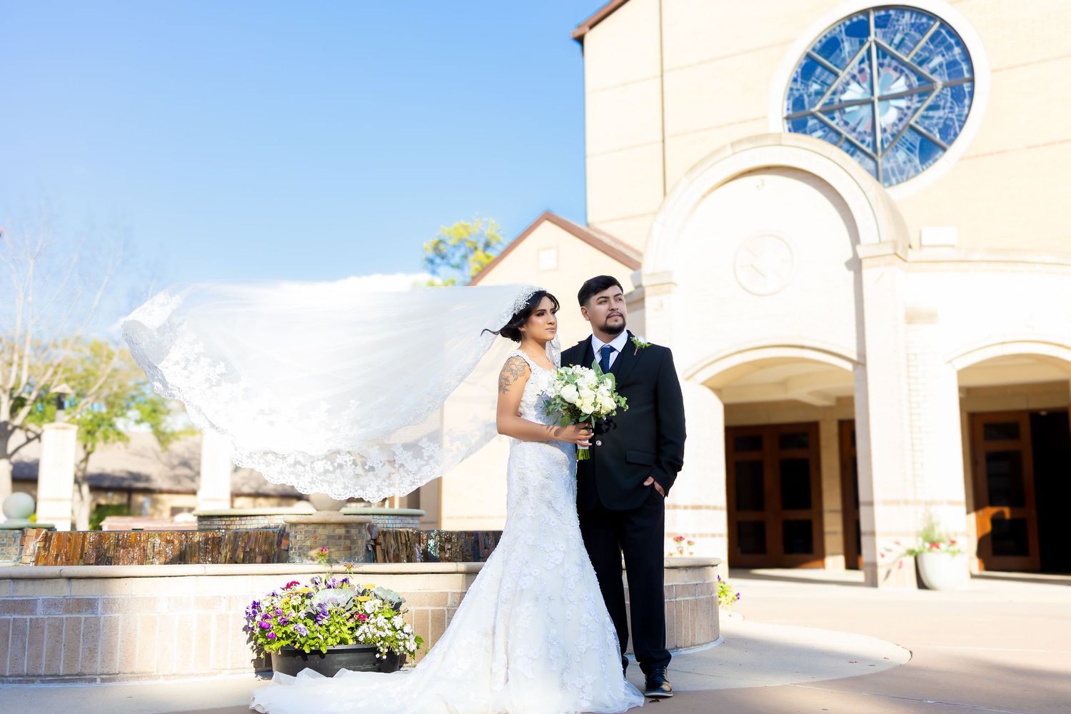 Exterior wedding photography at a white stucco church building under blue sky.