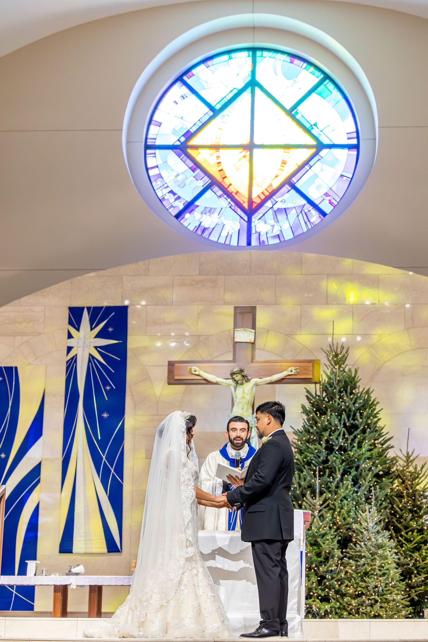 A wedding ceremony takes place in a modern church with stained glass windows and decorative Christmas trees.