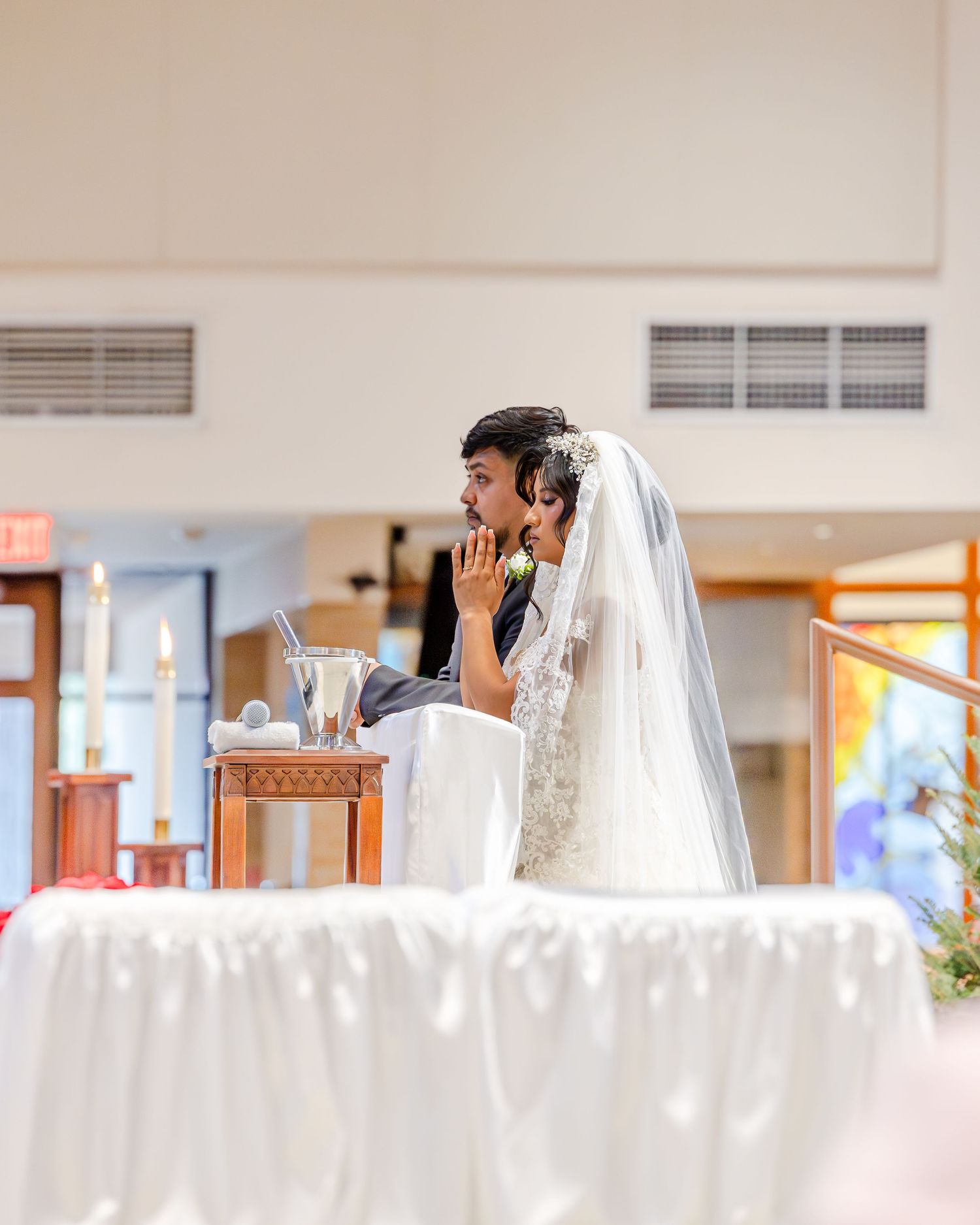 A bride in a white veil prays at a wooden altar during a church wedding ceremony.