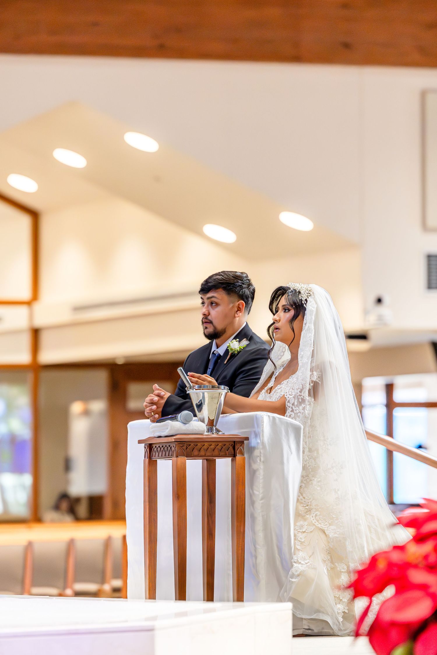 A bride and groom stand at a wooden podium during their wedding ceremony in a brightly lit church.