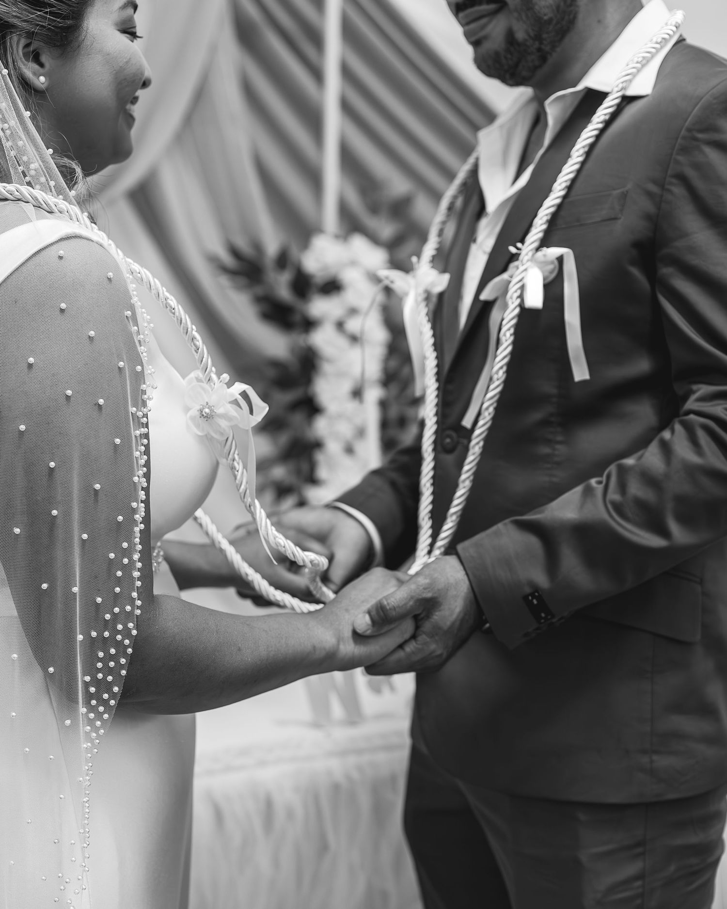 A couple in wedding attire holds hands during a traditional cord ceremony ritual at their wedding celebration.