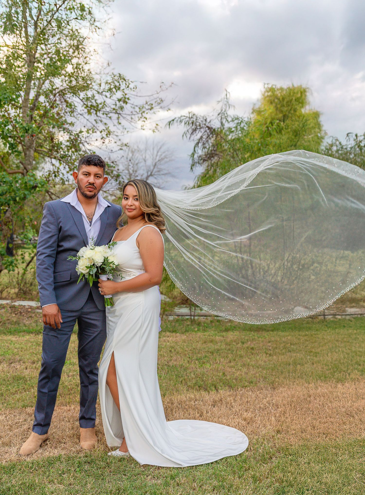 A romantic wedding couple stands in a field as a long white veil flows dramatically in the breeze.