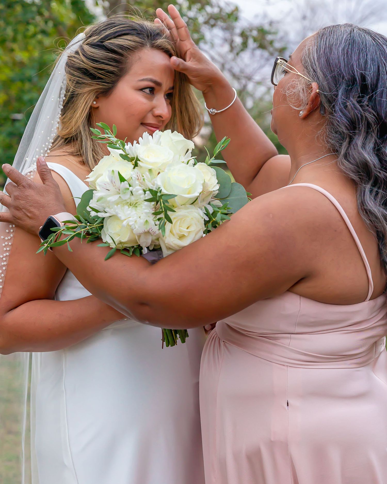 Bride and bridesmaid share an intimate moment while holding white floral bouquets at an outdoor wedding.