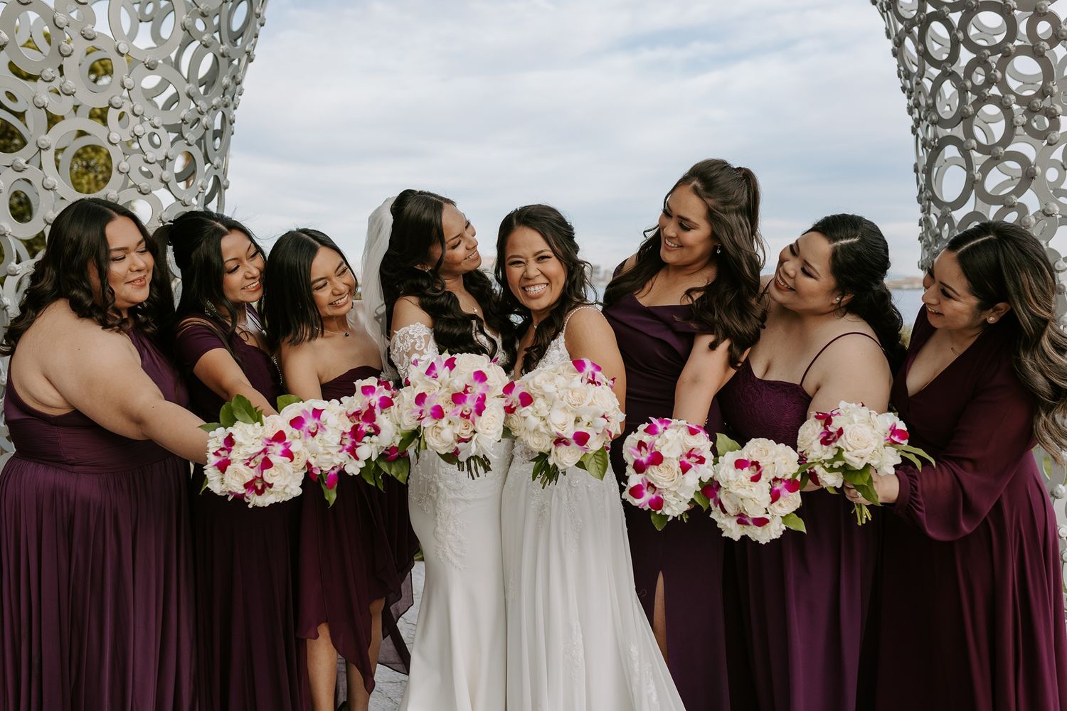 A group in dark purple and white dresses share joyful moments while holding pink and white floral bouquets at a wedding.
