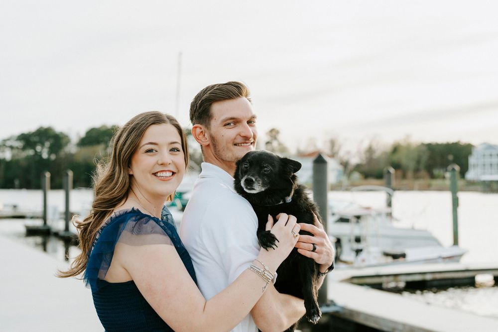 A couple stands at a marina holding a small black dog while smiling on a bright day.