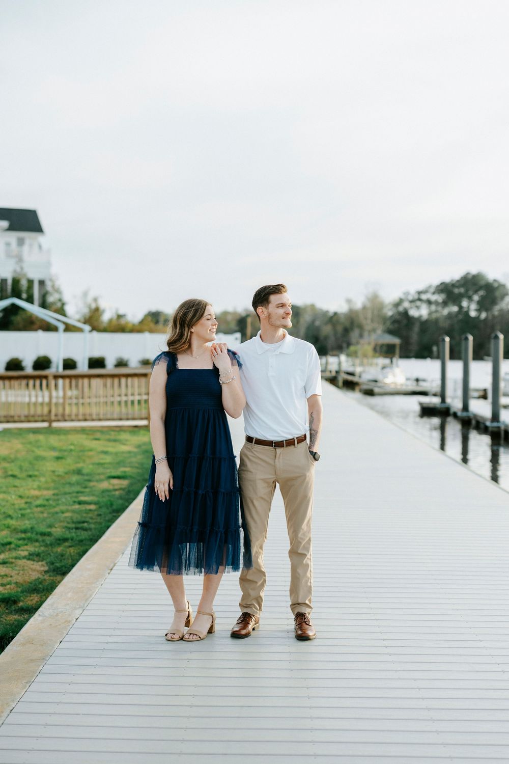 A couple walks together on a wooden boardwalk by the water on a sunny day.