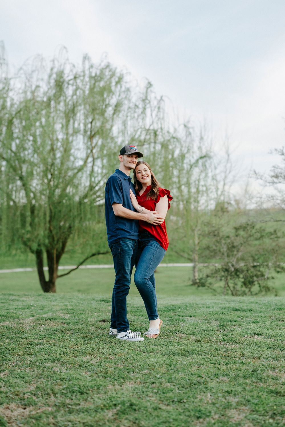 A couple shares playful moments on a grassy hillside with willow trees in the background.