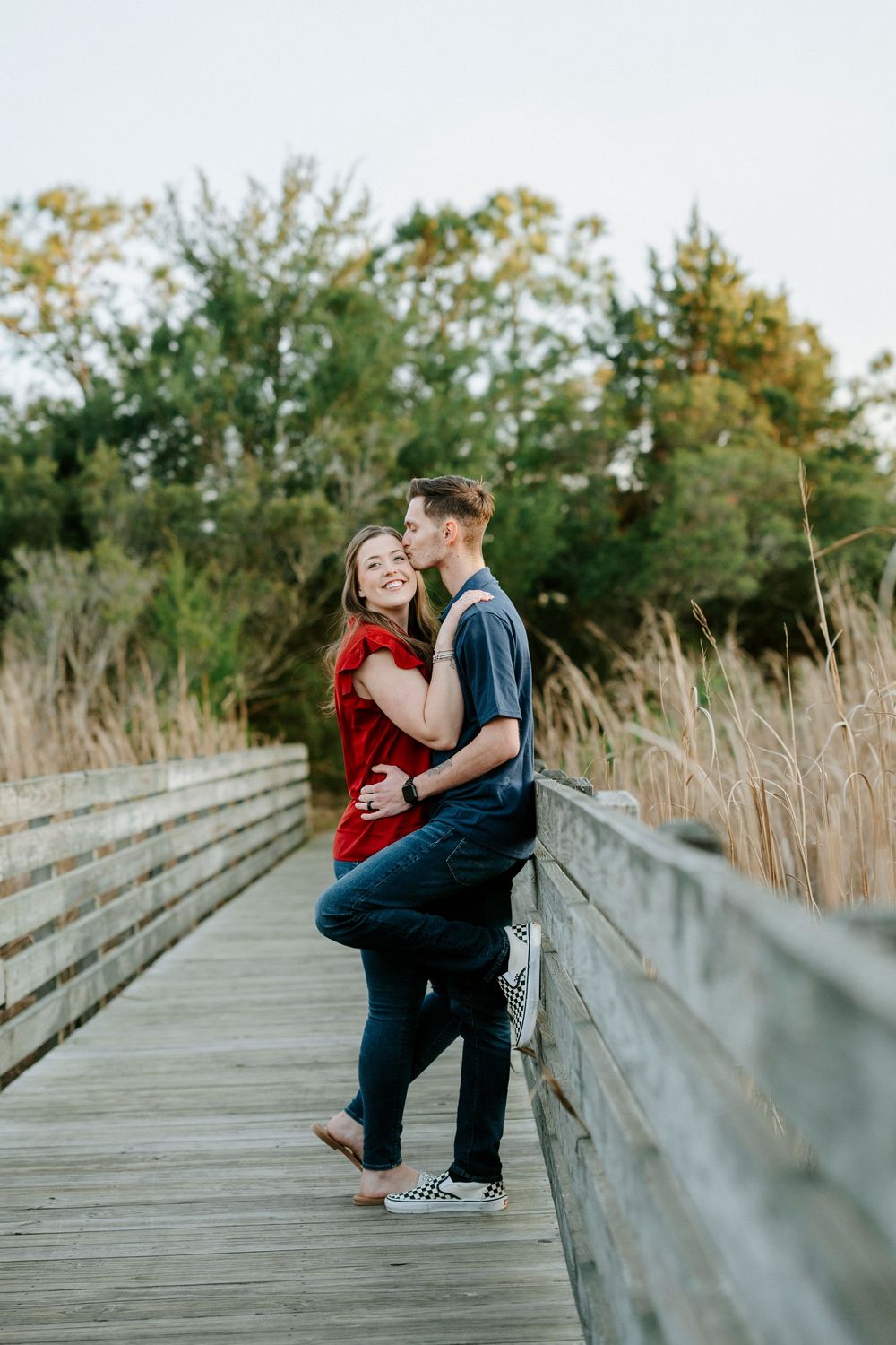A romantic couple shares intimate moments on a wooden boardwalk surrounded by autumn foliage and tall grasses.