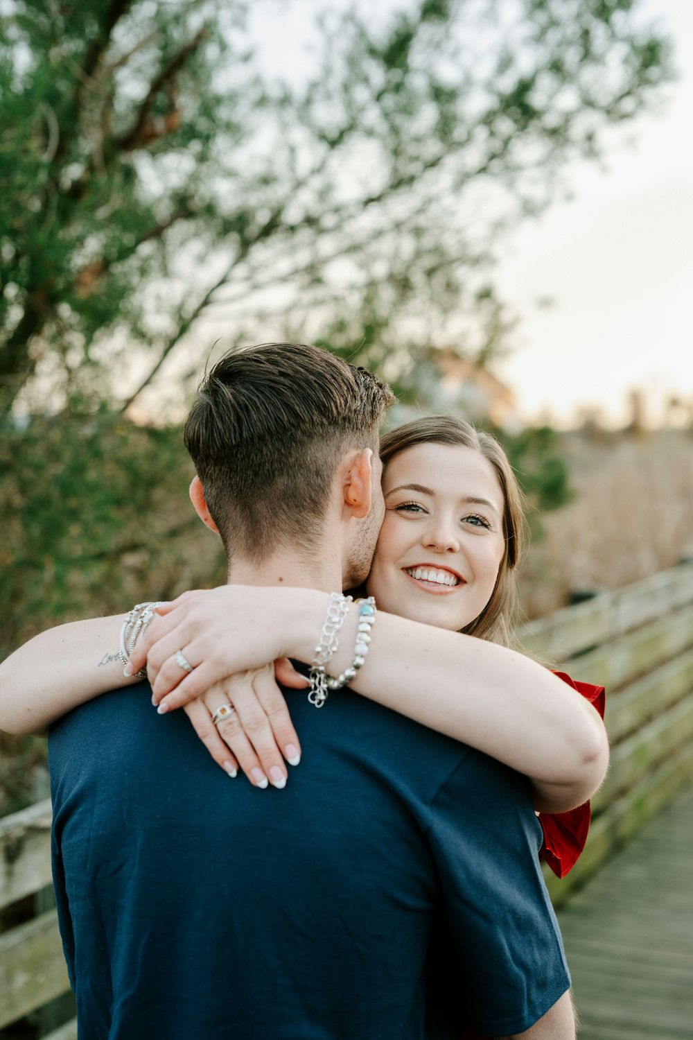 A couple embraces and smiles while standing outdoors with trees and greenery in the background.