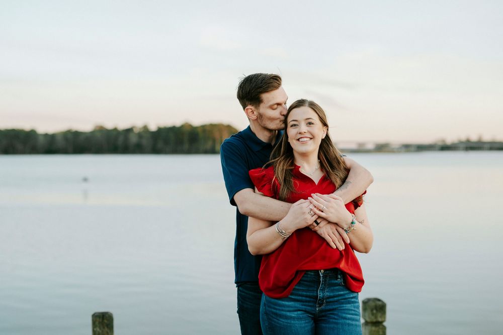 A couple in red and blue outfits embraces by a serene waterfront during a romantic photoshoot at sunset.
