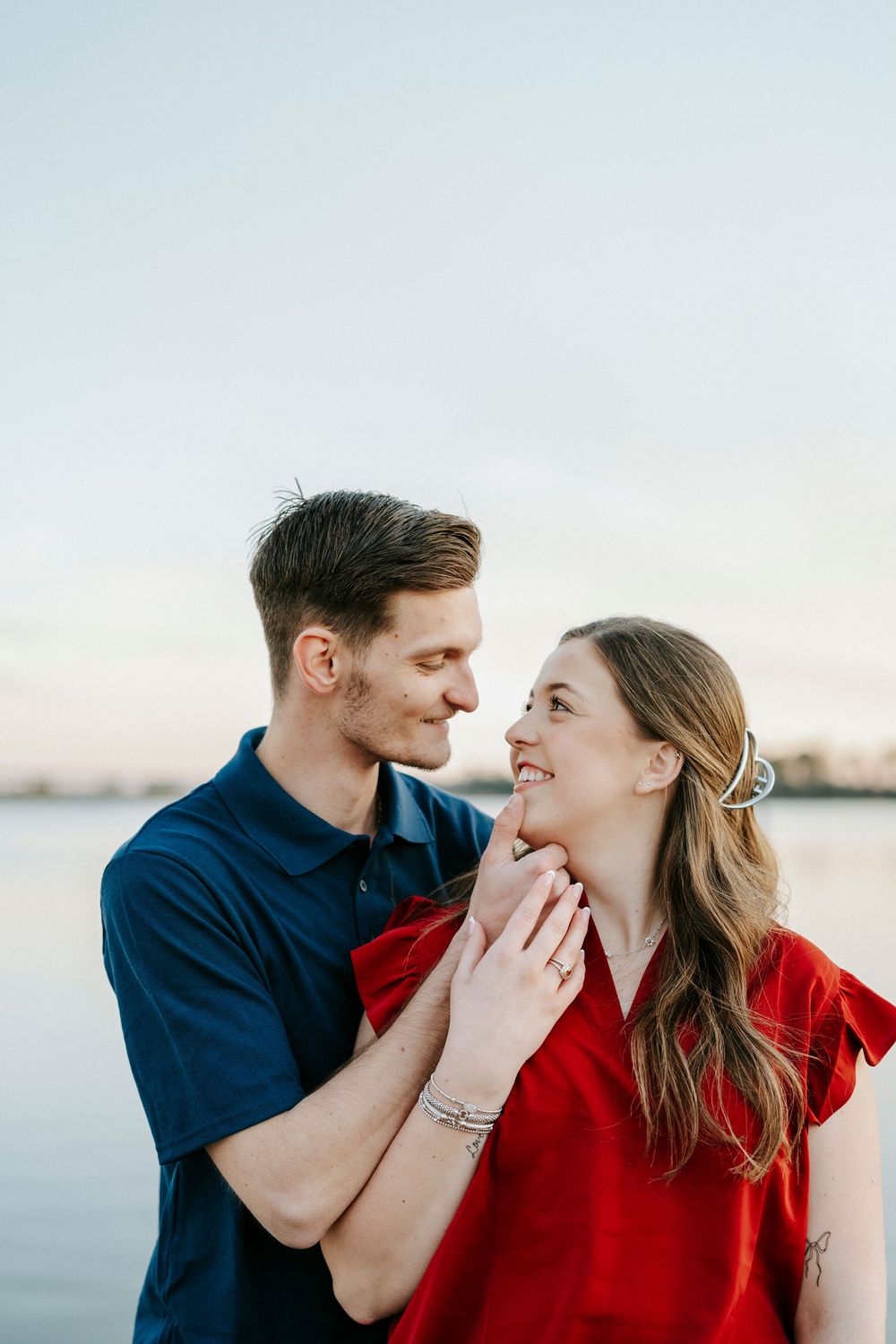 A couple in navy and red outfits share an intimate moment by the waterfront at sunset.