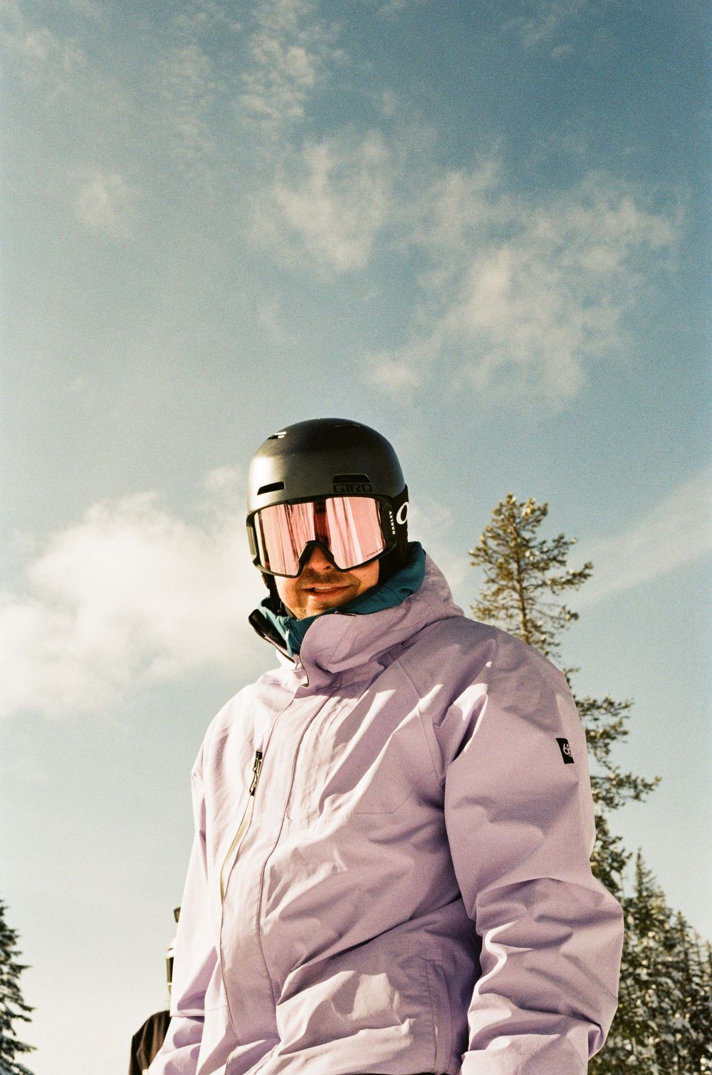 Snowboarder in lavender jacket and black helmet with pink mirrored goggles poses against a blue sky with snow-covered trees.