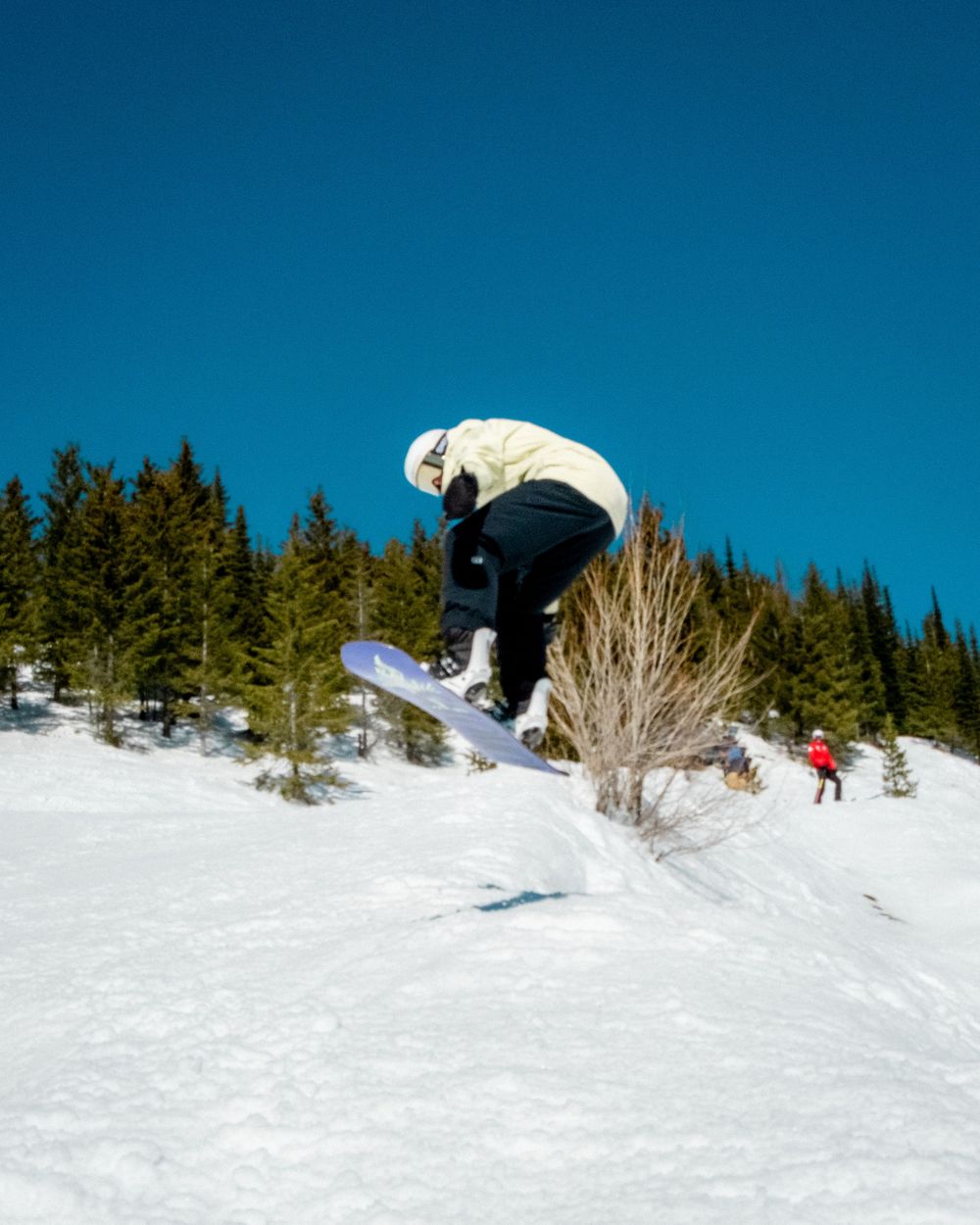 Snowboarder in white jacket performing aerial jump over snowy terrain with evergreen trees in background.