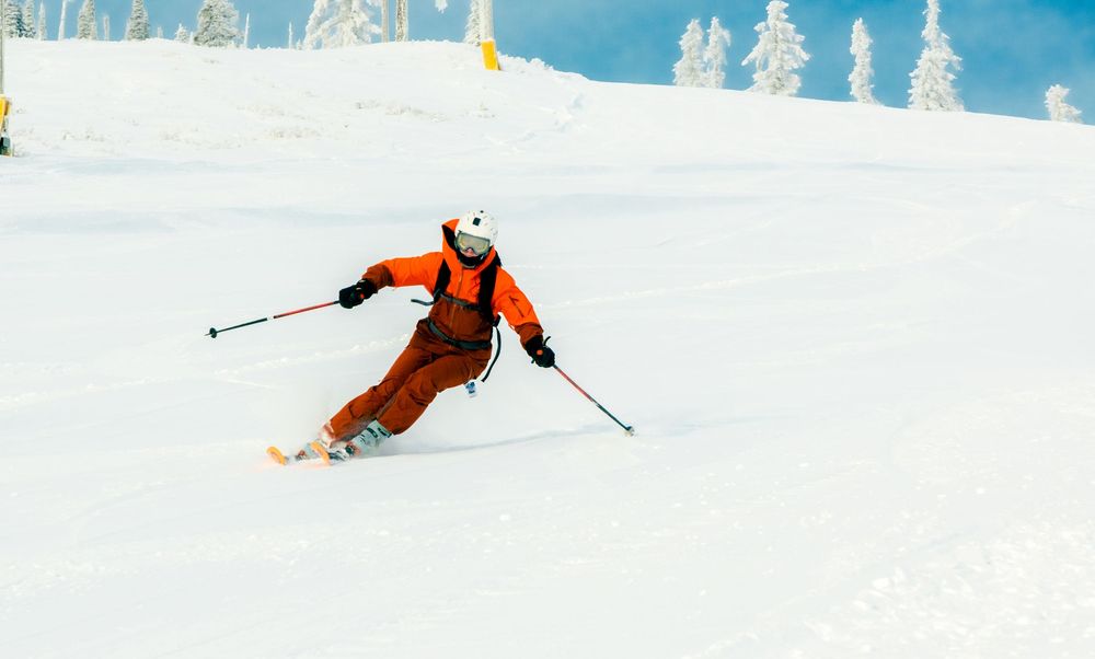 A skier in an orange jacket carves down a snowy slope with snow-covered evergreen trees in the background.