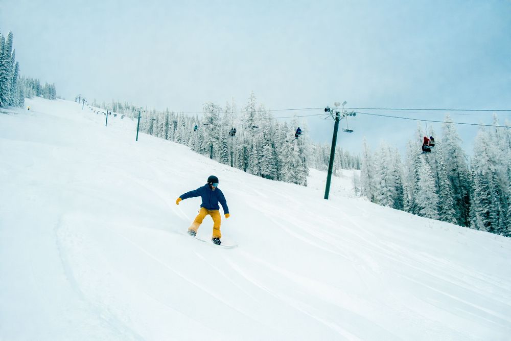 A snowboarder performs a jump against a snowy mountain backdrop with ski lifts visible in the distance.