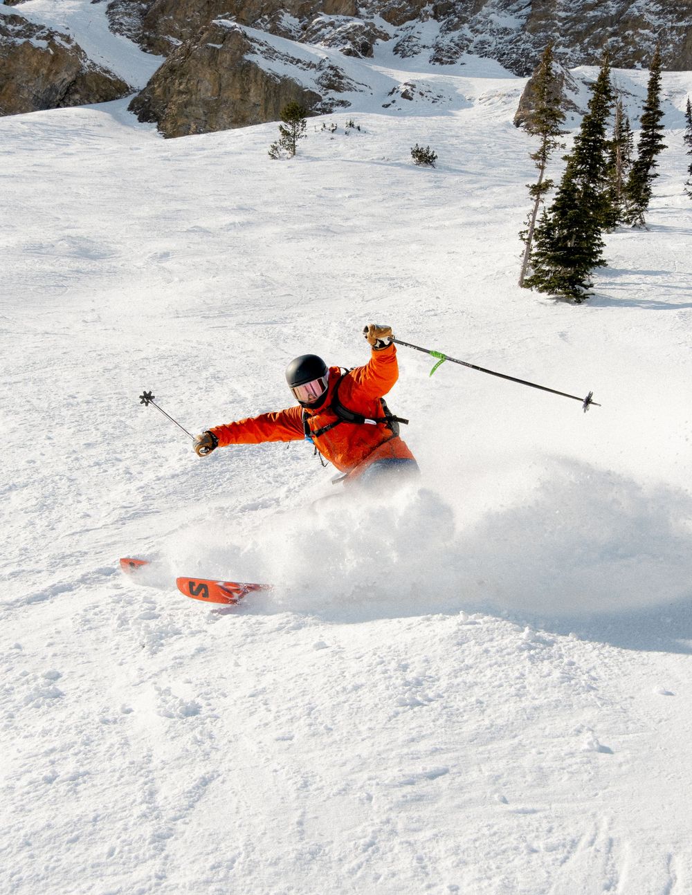 A skier in bright orange gear carves through deep powder snow on a mountain slope, creating a dramatic spray of snow.