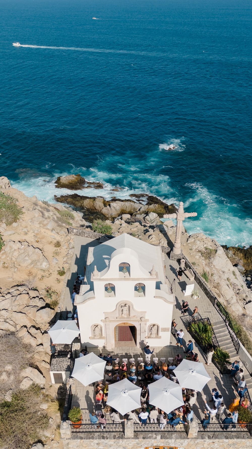 Aerial view of a white chapel with outdoor dining area perched on rocky cliffs above deep blue ocean.