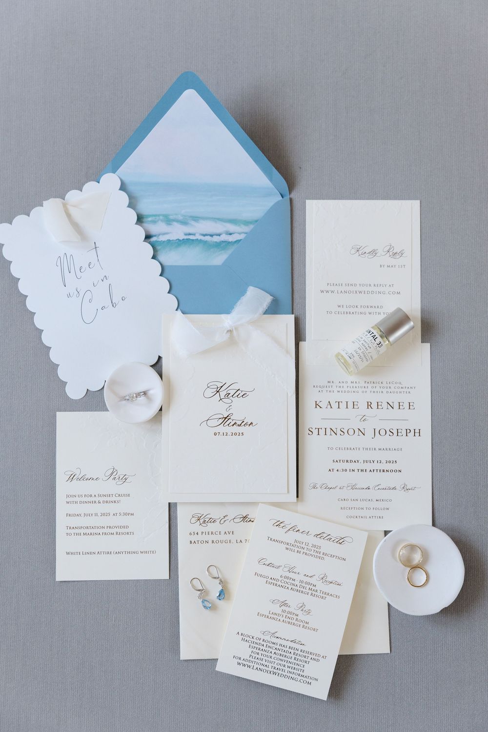 Elegant wedding invitation suite with ocean-themed blue envelope liner, rings, and earrings on gray background.