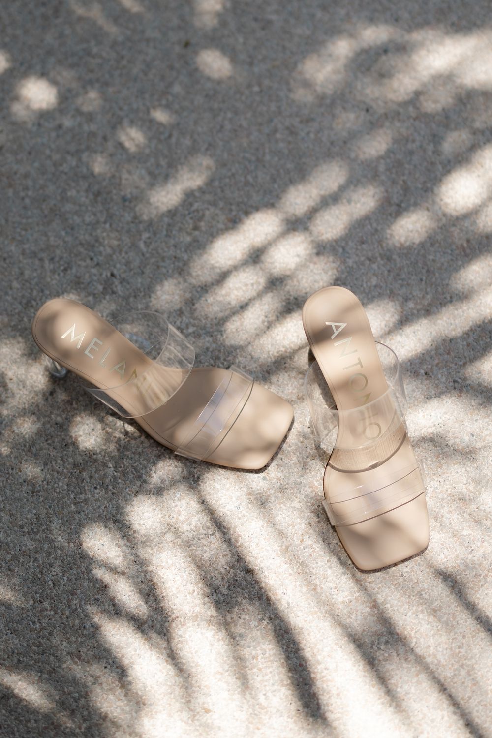Neutral beige clear-strap heeled sandals on sunlit concrete with dappled shadow patterns.