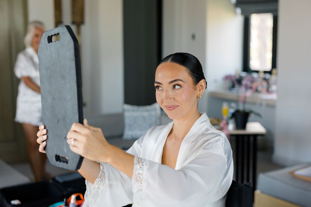 Bride in white lace robe checking her look in a handheld mirror while getting ready for her wedding.