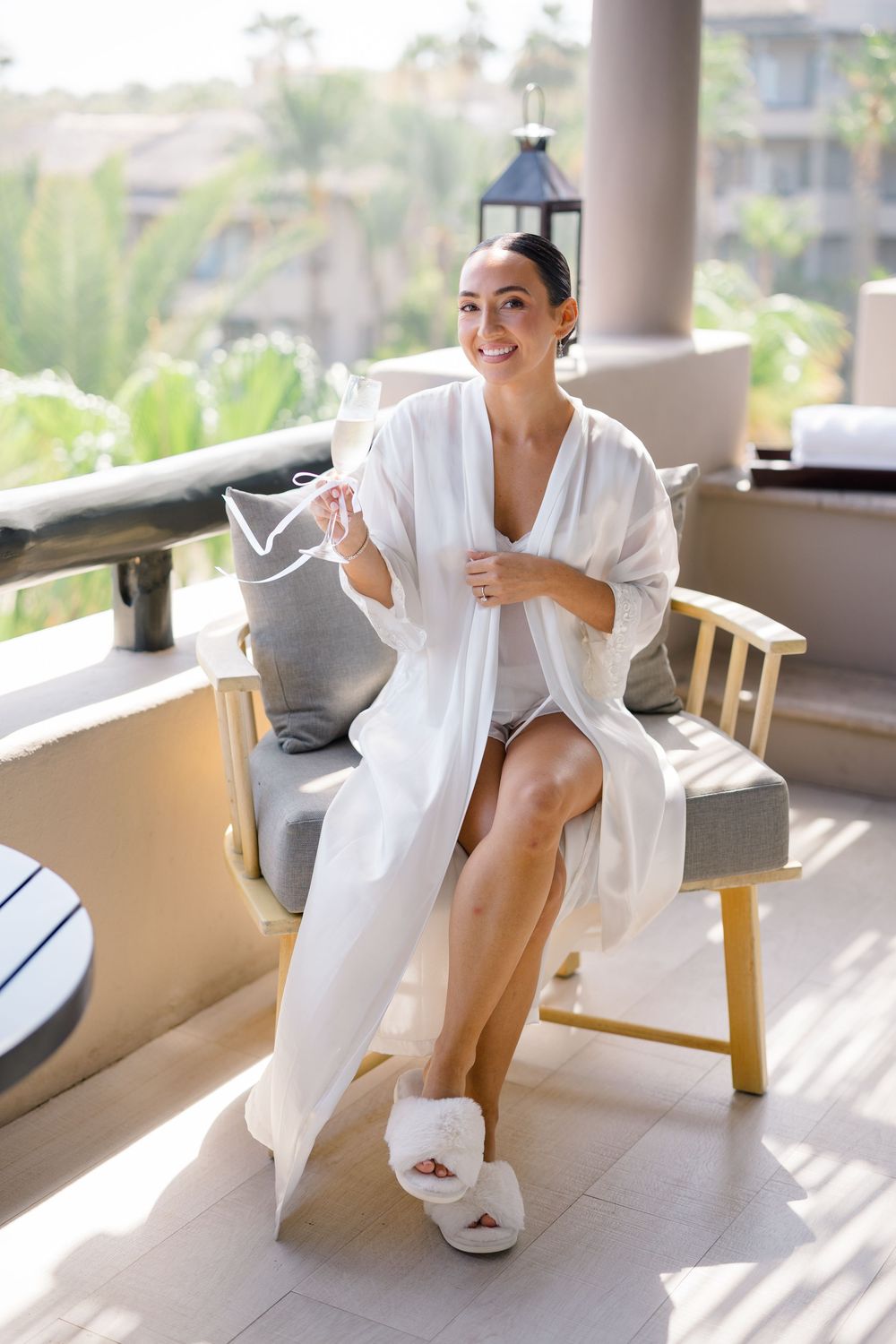 Smiling bride in white robe and slippers relaxing on a balcony with champagne surrounded by palm trees.