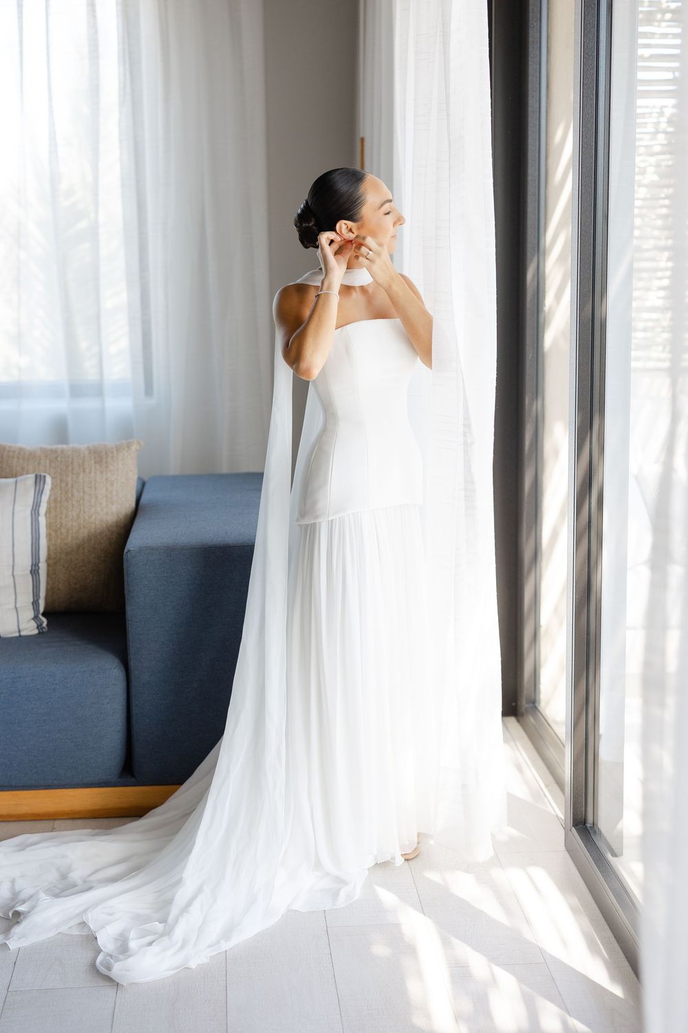 Elegant bride in strapless white gown with long cathedral veil putting on earrings by a sunlit window.