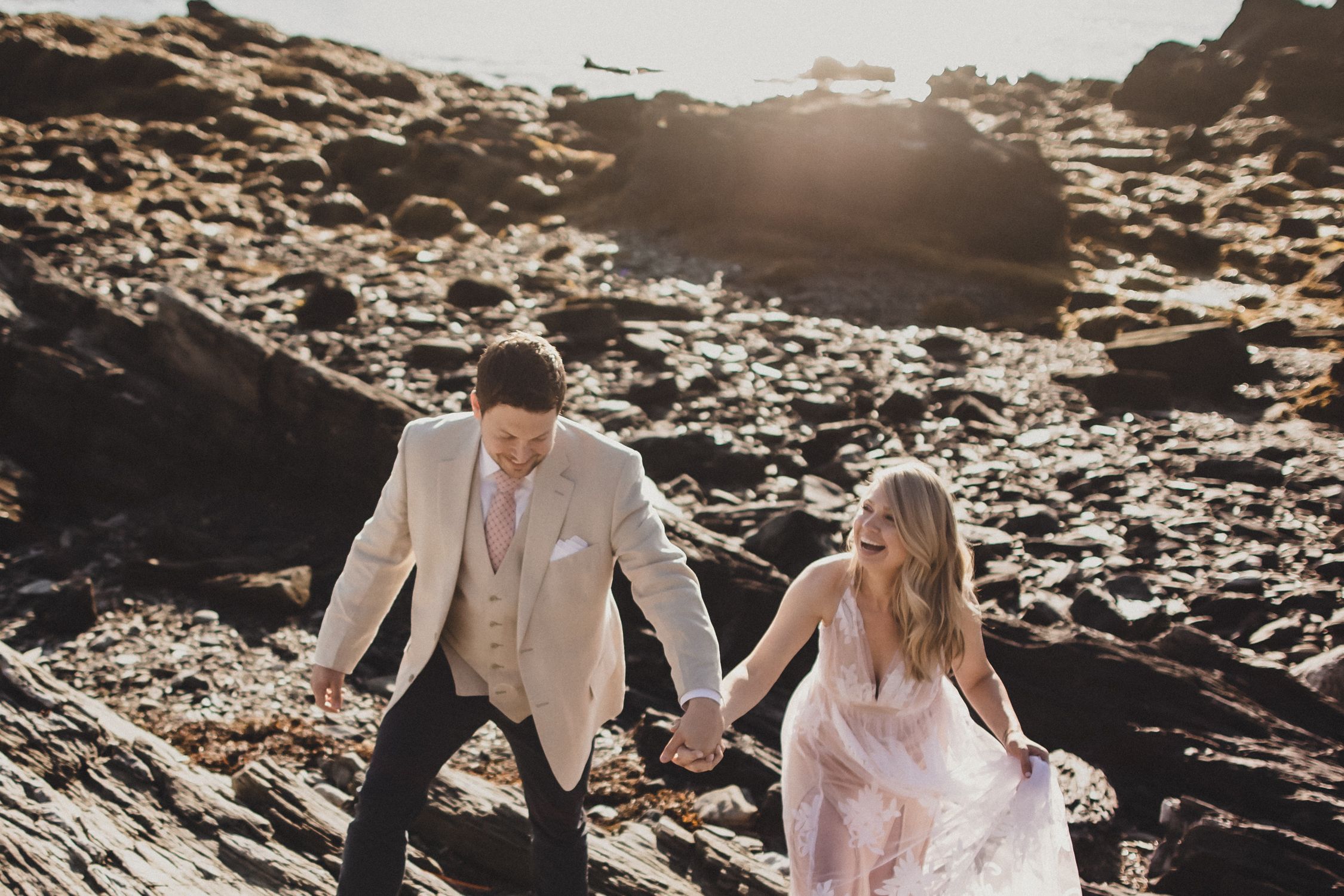 Couple in wedding attire shares romantic moments on dramatic rocky coastline at sunset.
