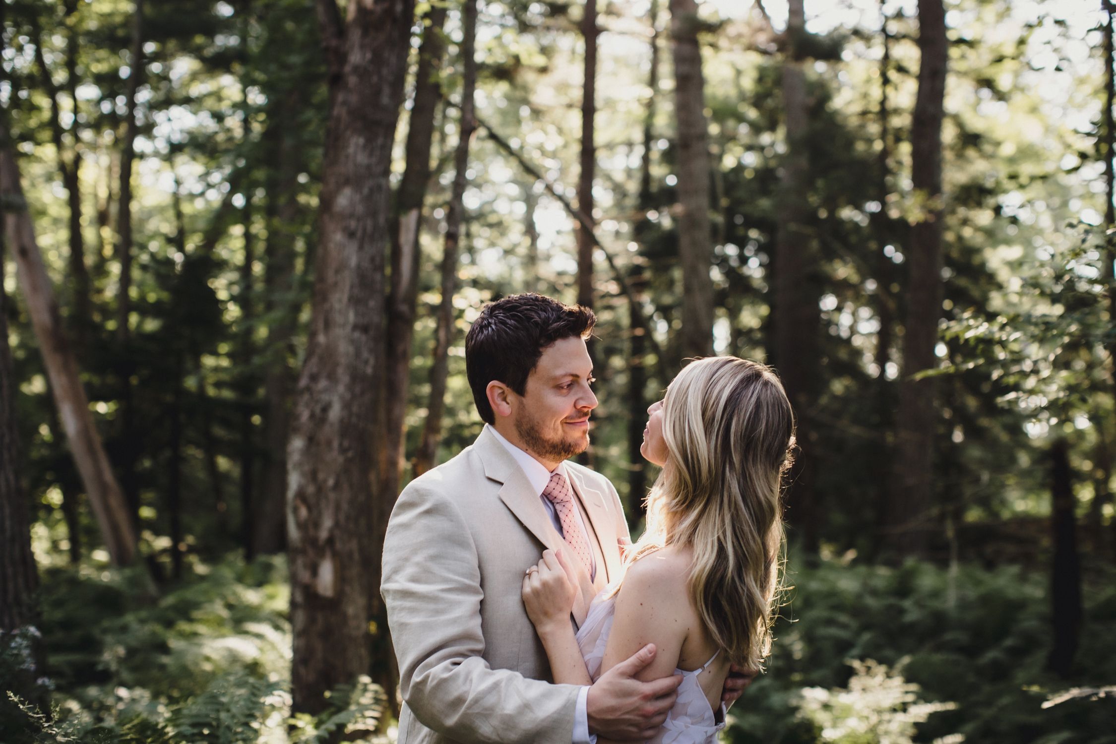 A couple in formal attire shares an intimate moment together in a sunlit forest setting.