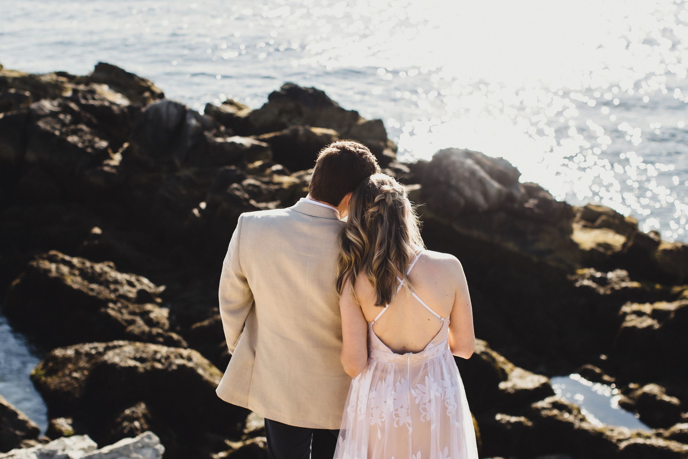 A couple in formal attire stands together overlooking a rocky ocean coastline at sunset.