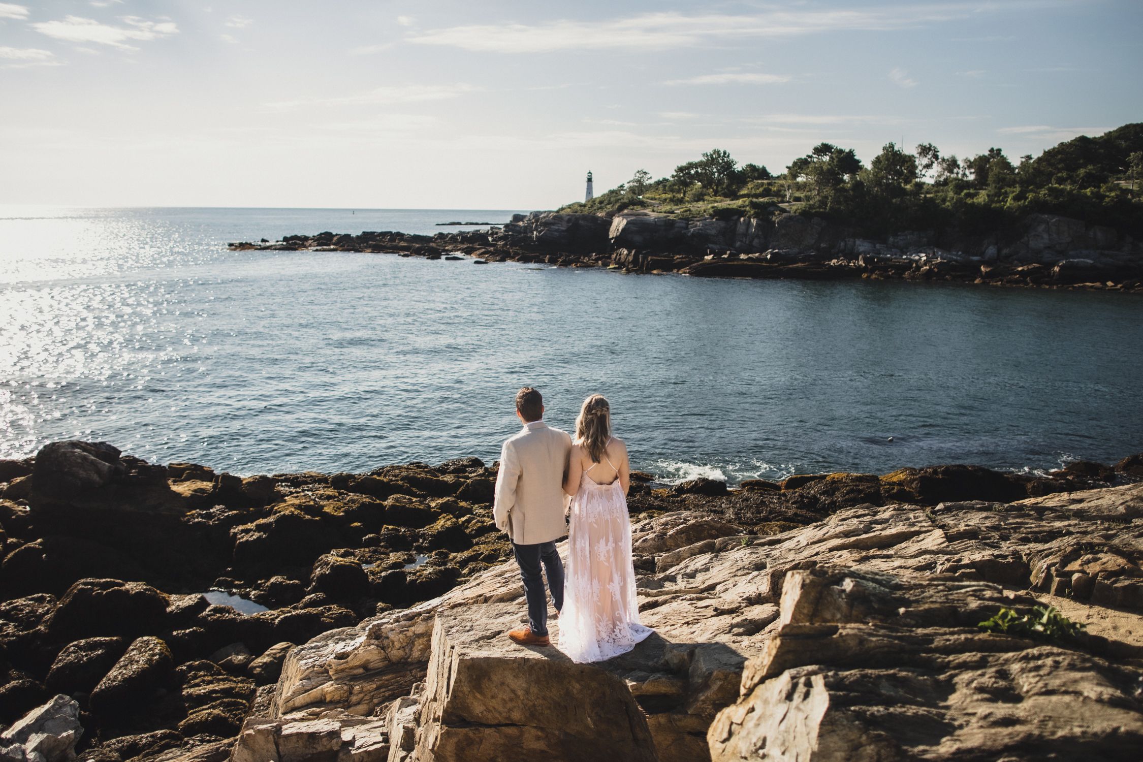 Couple in wedding attire embracing on rocky coastal cliffs with sparkling ocean and blue sky in background.