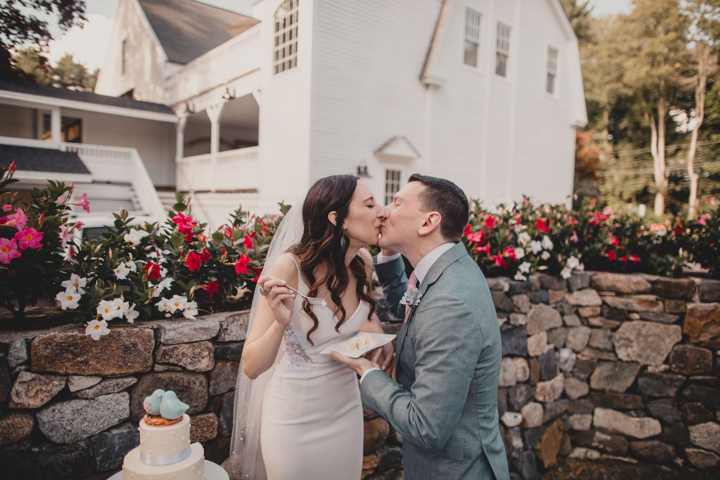 Couple embracing in front of a rustic stone wall with blooming red roses in an outdoor garden setting.