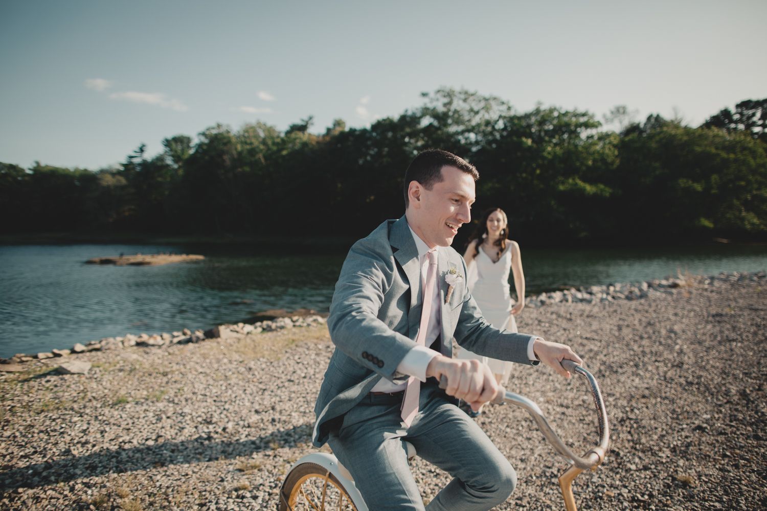 A couple enjoying a sunny day by a rocky lakeshore as someone rides a bicycle in a gray suit.