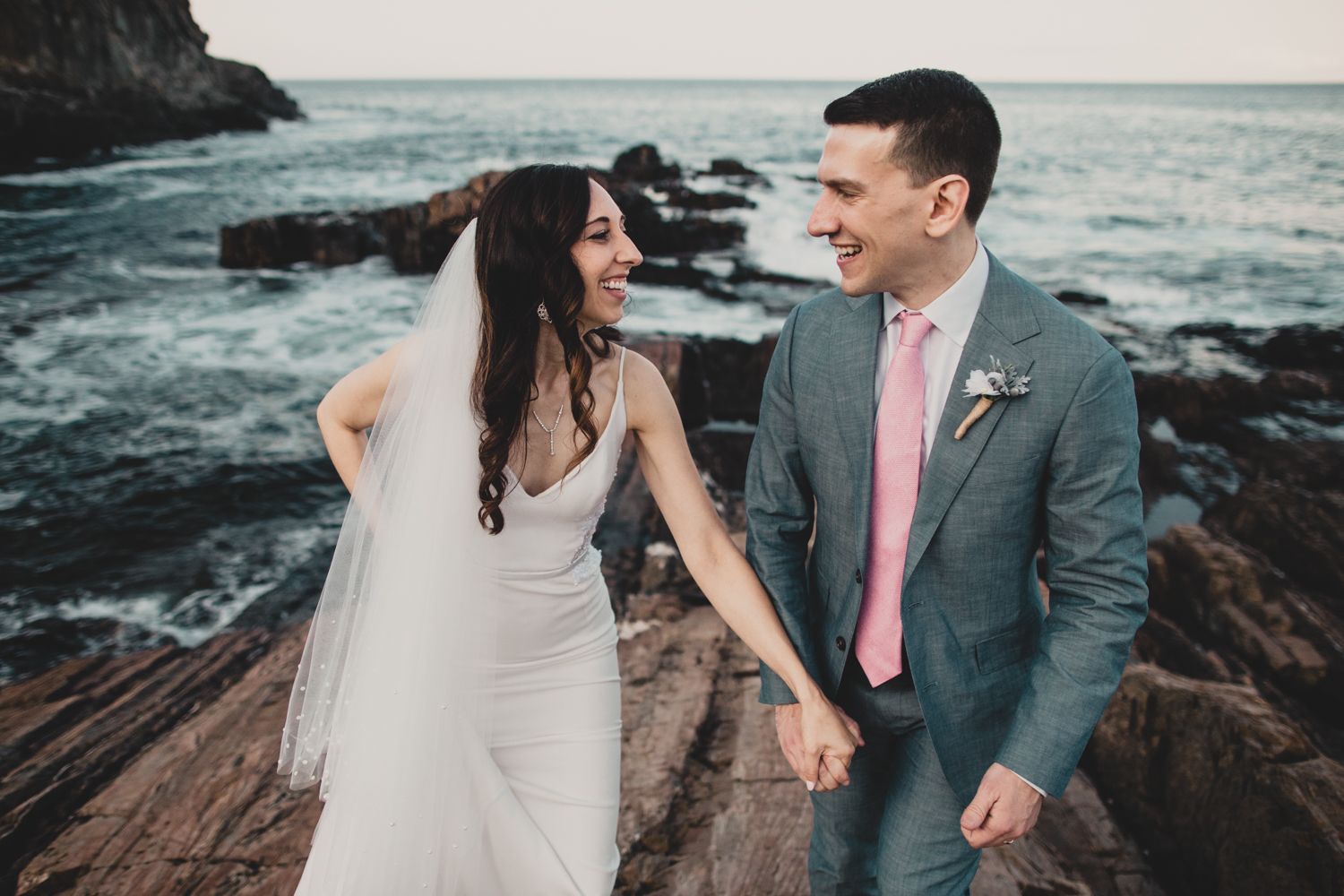 A romantic wedding couple stands on rocky coastal cliffs with crashing waves in the background at sunset.