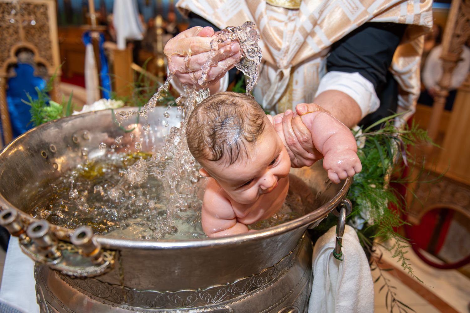 Priest performing Orthodox Christian baby baptism ceremony, immersing infant in decorative silver font.