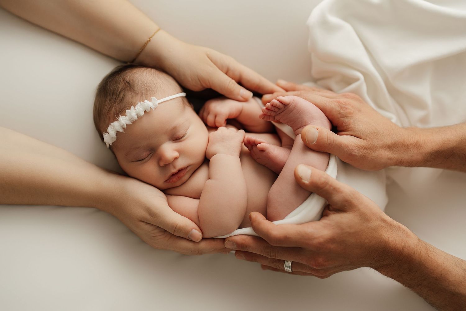 A sleeping newborn wearing a delicate floral crown lies peacefully on soft white textured fabric in a studio setting.