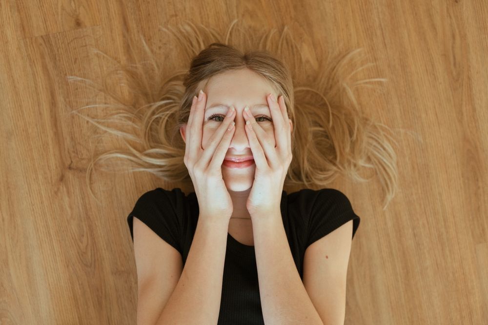 A person with blonde hair lying on wooden floor covers their face playfully while laughing.
