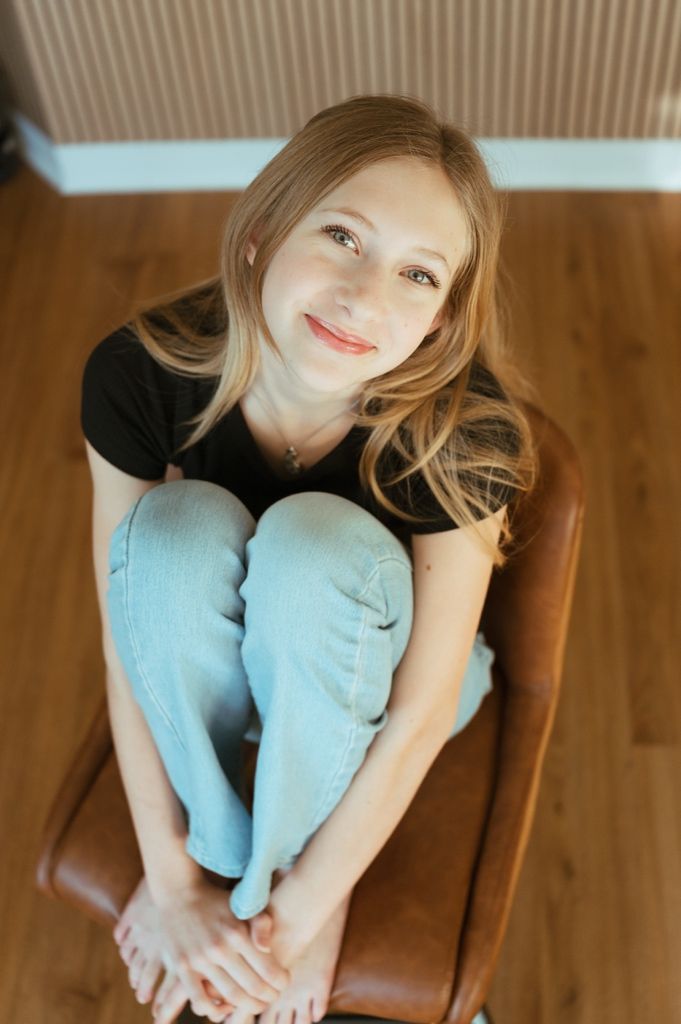 A casual portrait of someone sitting on a brown leather chair wearing light denim and black top.