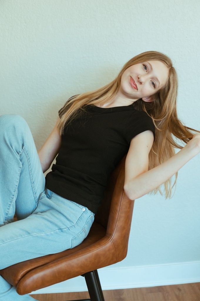 Fashion model in black dress reclining on brown leather chair against light blue wall.