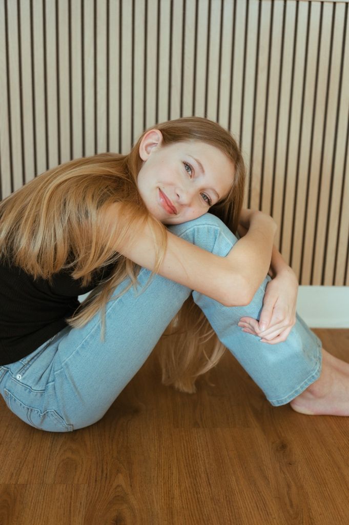 A person in light blue jeans and black top sits casually against a wooden paneled wall.