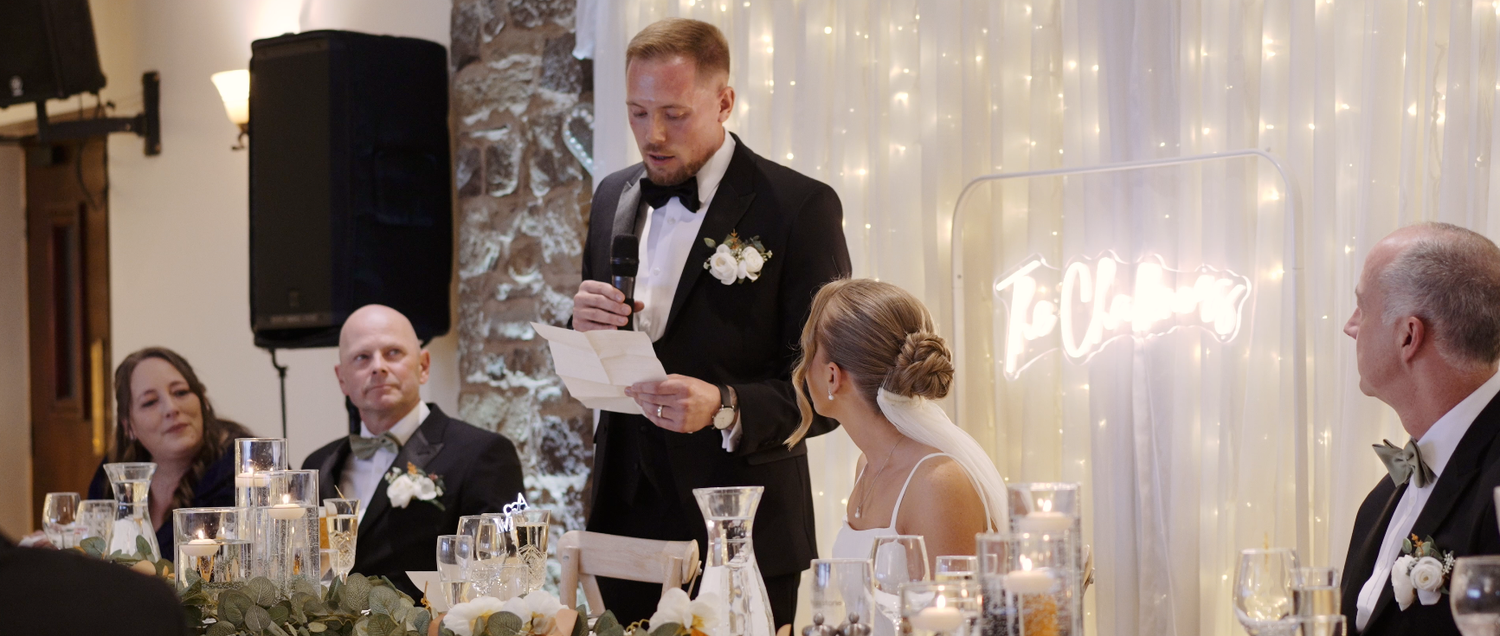 Wedding reception guests seated at elegant tables with white decor and ambient lighting during a dinner toast.