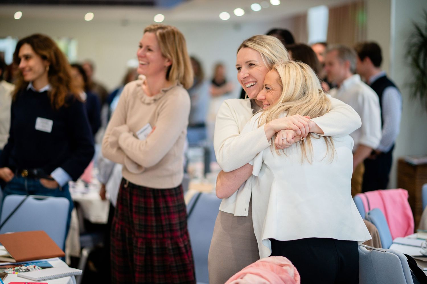 Two women share a warm hug at a professional networking event, smiling joyfully, with other attendees visible in the background.